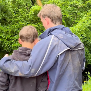 two young men seen from the back, the taller has a friendly arm round the other's shoulder
