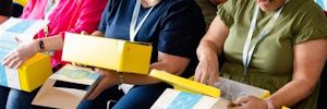 two lines of people with yellow cardboard boxes on their laps smiling and opening them up